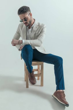 sexy cool businessman with retro sunglasses looking down and posing while sitting on wooden chair, holding arms in fashion pose on grey background