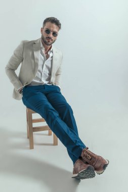 handsome bearded man with hand in pockets wearing retro glasses and sitting on wooden chair on grey background 