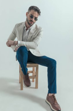 attractive young man in suit with undone shirt holding arms in a fashion pose while sitting on wooden chair on grey background in studio