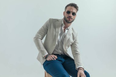 Takım elbiseli yakışıklı genç bir işadamı ahşap koltukta oturmuş, moda pozunda el ele tutuşuyor ve gri arka planda poz veriyor.