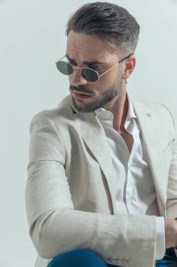 havalı traşsız iş adamı, retro güneş gözlüklü dirseğini dizine dayamış ve gri arka planda otururken ve poz verirken yana bakıyor.