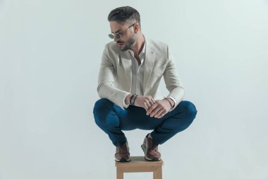 attractive crouched man with sunglasses touching palms and looking to side on grey background in studio