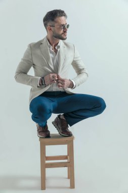 crouched bearded man looking to side and buttoning jacket while posing in front of grey background