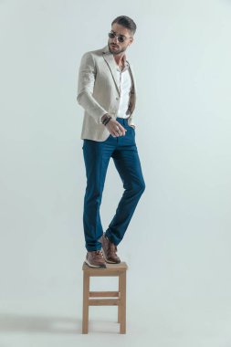 sexy man with open collar shirt holding hand in pocket and looking to side while standing on wooden chair on grey background 