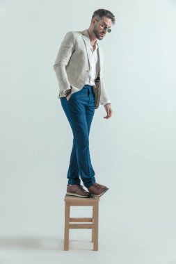 unshaved businessman holding hand in pockets and thinking of jumping into new life while standing on wooden chair in front of grey background