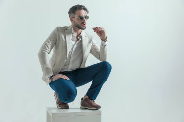 sexy businessman with open collar shirt looking to side and crouching, holding elbows on knees and posing on grey background