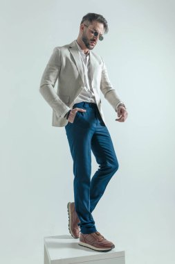 cool man with open collar shirt holding hand in pocket and looking down, posing in a confident manner on grey background in studio