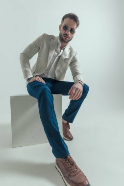 full body picture of cool bearded guy with glasses holding elbow on knees and sitting on grey background 