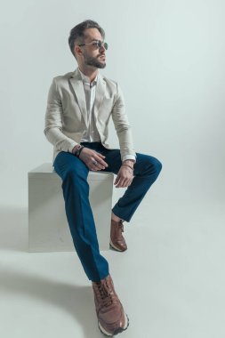 full body picture of sexy man with beard wearing glasses looking to side and sitting on grey background in studio