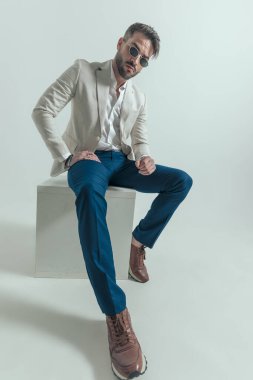 sexy unshaved businessman with open collar holding arms in a fashion pose, sitting on grey background in studio and posing
