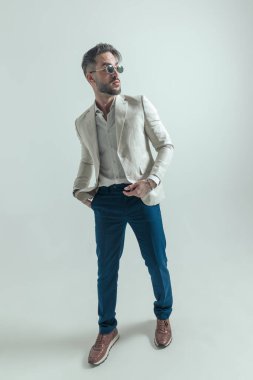 full body picture of bearded man with hand in pocket looking to side and posing in a cool and fashion manner on grey background in studio