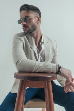 Gözlüklü seksi bir adam ve kolları bağlı olmayan, tahta koltukta oturan ve stüdyoda gri arka planı olan bir adam.