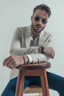 Gözlüklü havalı bir iş adamı, tahta koltukta dirseklerini tutuyor ve kendine güveni tam, gri arka planda moda bir şekilde poz veriyor.