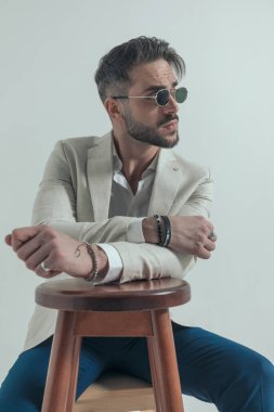 Stüdyoda oturan sakallı iş adamı, gri arka planda otururken ahşap koltukta durup kollarını tutuyor.