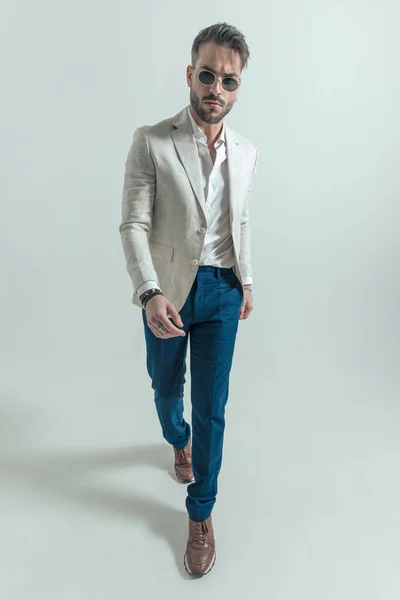 sexy man with open collar wearing sunglasses and confidently walking on grey background in studio