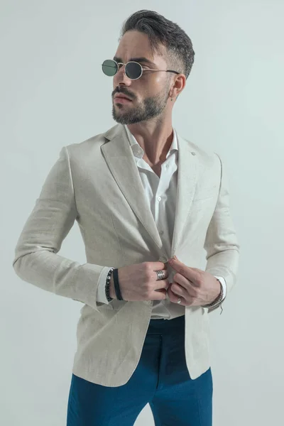 confident man with sunglasses and open collar shirt adjusting jacket and looking to side on grey background in studio