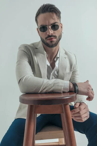 木製の椅子に腕を保持し 灰色の背景にクールな態度でポーズを取るオープン襟シャツを持つ魅力的なビジネスマン — ストック写真