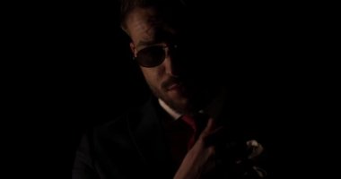 Güneş gözlüklü gizemli bir genç adam sakala dokunuyor ve ovuyor, yanına bakıyor ve karanlıkta gülümsüyor