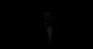 Smokinli yakışıklı adam kollarını kavuşturuyor, gülümsüyor ve siyah arka planda poz veriyor.
