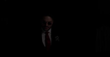 elegant man in suit with tie and sunglasses coming from the dark, looking to side, crossing arms, sensually touching lips on black background