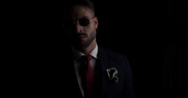 cool sexy businessman looking to side, adjusting tie and sunglasses and confidently folding arms on black background in the dark