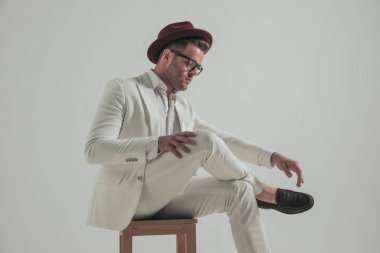 Beyaz takım elbiseli, bacak bacak üstüne atmış, ahşap sandalyede otururken kendine güvenen havalı bir adamın yan görüntüsü. 