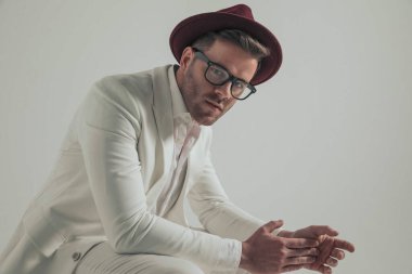 side view of seated fashion businessman in white suit with hat touching palm with finger and posing on grey background in studio