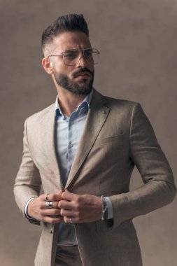 Seksi sakallı iş adamı takım elbise ve düğme arıyor. Stüdyoda bej arka planda havalı ve kendinden emin.
