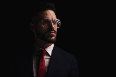 Gözlüklü, havalı, tıraşsız bir iş adamı stüdyoda siyah arka planda poz veriyor.