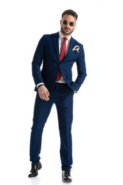 Deniz mavisi takım elbiseli, gözlüklü, kendine güveni tam bir sakallı adam. Cebinde gözlükleriyle stüdyoda beyaz arka planda poz veriyor.