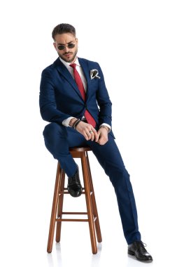 Takım elbiseli genç adam ahşap koltukta oturuyor, stüdyoda beyaz arka planda havalı bir poz veriyor.