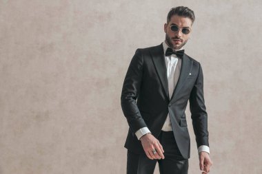 sexy bearded businessman in black tuxedo with sunglasses posing in front of beige background in studio