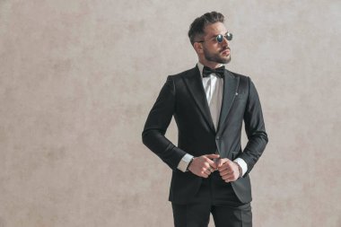 attractive elegant man looking to side while adjusting black tuxedo, being confident and cool, posing and standing on beige background