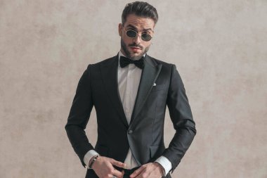 portrait of elegant businessman in black tuxedo wearing sunglasses and posing in front of beige background in studio