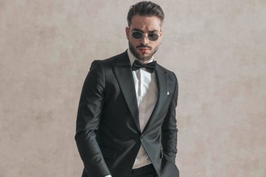 portrait of sexy bearded businessman with sunglasses and hand in pocket posing in front of beige background in studio