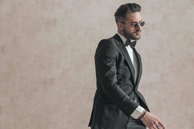 Siyah smokinli seksi damadın yandan görünüşü moda pozunda el ele tutuşması ve stüdyoda bej arka planda havalı bir poz vermesi.