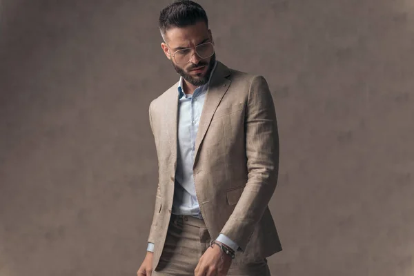 sexy elegant man with open collar shirt looking down and posing while standing in front of beige background in studio