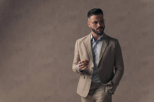 cool fashion businessman in suit with open collar shirt, with one hand in pocket, holding the other hand in a fashion pose while looking to side on beige background