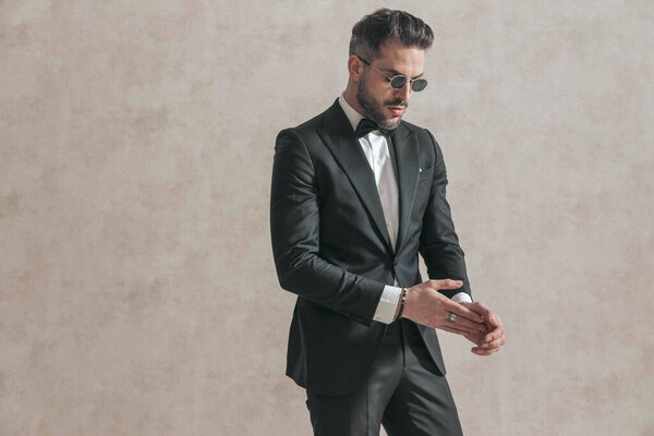 sexy elegant man wearing black tuxedo with bowtie rubbing palms and being confident in front of beige background in studio
