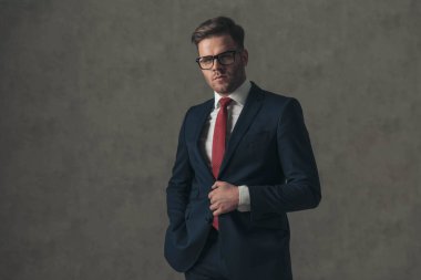 sexy young businessman with eyeglasses unbuttoning elegant suit, holding hand in pocket and posing in front of beige background in studio