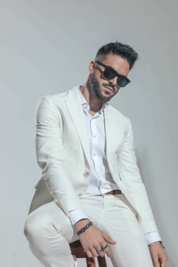 sexy stylish man in white suit with sunglasses posing in a cool way on grey background in studio 