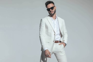 smart casual guy in white suit with open collar shirt holding hand in pocket and confidently posing on grey background 