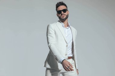 portrait of unshaved cool man wearing shite stylish suit holding arms in fashion pose, being confident and posing in front of grey background