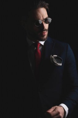bearded man in elegant suit with red tie and retro sunglasses, wearing handkerchief in the pocket and looking to side while posing