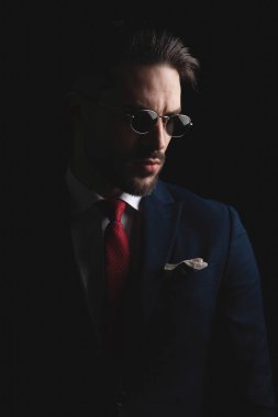 sexy elegant man in suit with red tie and retro sunglasses looking to side and posing in a dramatic way on black background in studio