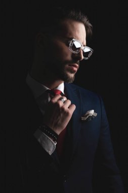 portrait of attractive elegant man in suit adjusting red tie and posing in a cool way while looking to side on black background