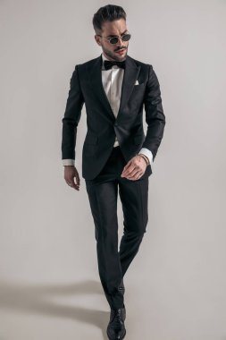 confident young groom in tuxedo with glasses looking away and walking on grey background in studio