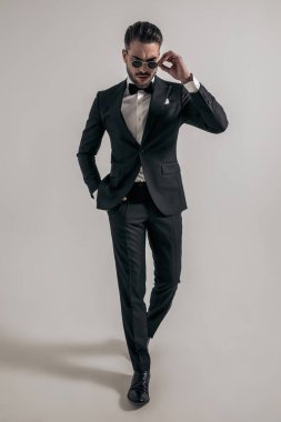 full length picture of cool elegant man in tuxedo looking down and fixing sunglasses while walking with hand in pocket on grey background