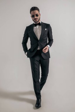 bearded young groom with hand in pocket wearing elegant tux and sunglasses and walking on grey background in studio