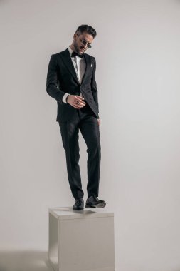 handsome young groom in tuxedo with sunglasses looking down and stepping out of wooden box in front of grey background